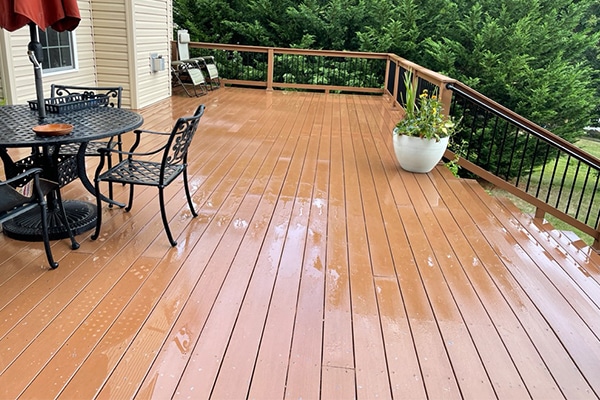 Wet, brown deck with black metal patio furniture and potted plants. The deck overlooks a lush green yard and trees.