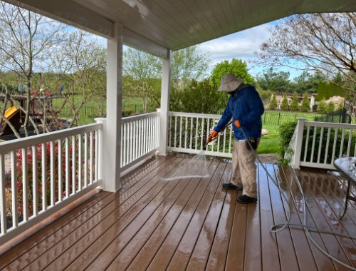 A person power washing a wood deck on a sunny day, wearing a hat and blue jacket, with lush green lawn and trees in the background. Power washing maintenance.