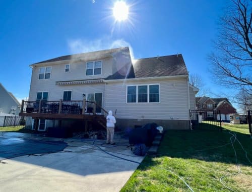 A person power washing the siding of a two-story house on a sunny day. Water spray is visible as they clean, with a green lawn and blue sky in the background.