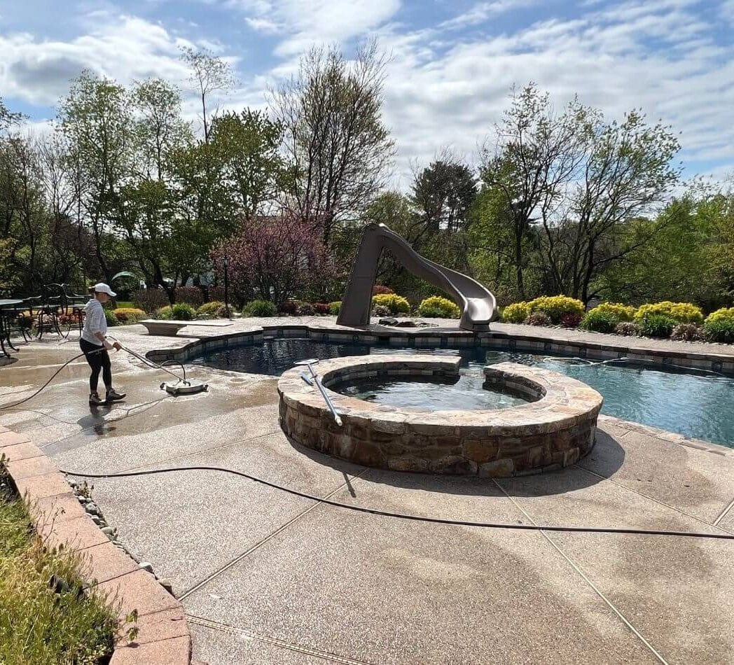 Person power-washing a concrete pool deck beside a round stone spa and slide, backyard pool with trees and shrubs.