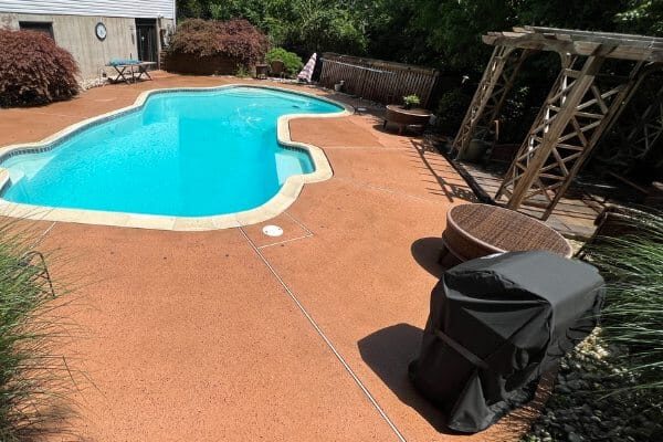 Backyard kidney-shaped pool with clear blue water on red-orange concrete deck, covered grill, wicker seating and wooden pergola.
