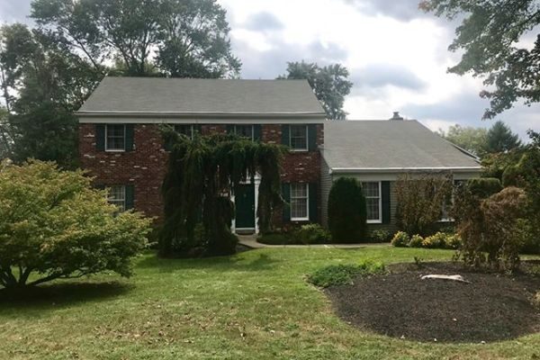 Two-story red brick colonial house with attached one-story wing, front lawn, shrubs and a weeping tree over the walkway under a cloudy sky.
