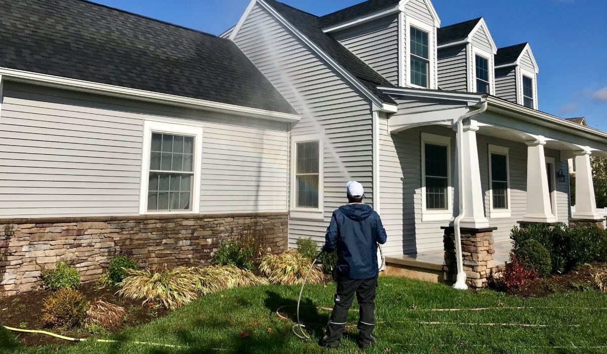 Person in jacket and cap pressure-washing the siding of a two-story house, water spray arching over the lawn and shrub beds.