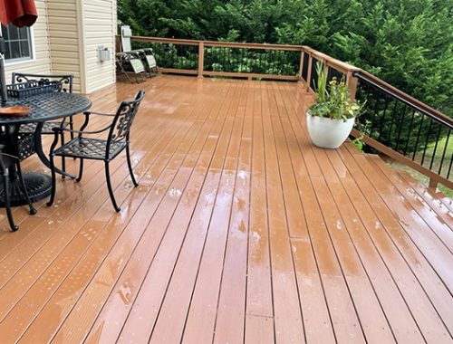 Wet, brown deck with black metal patio furniture and potted plants. The deck overlooks a lush green yard and trees.
