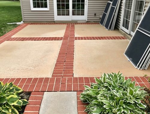 Outdoor patio with red brick border and beige concrete squares. Screen doors lean against the house, with green hosta plants in the foreground.