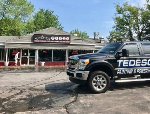 Black pickup labeled TEDESCO Painting & Powerwashing parked before Kosmos Pizza storefront on cracked asphalt, trees behind.