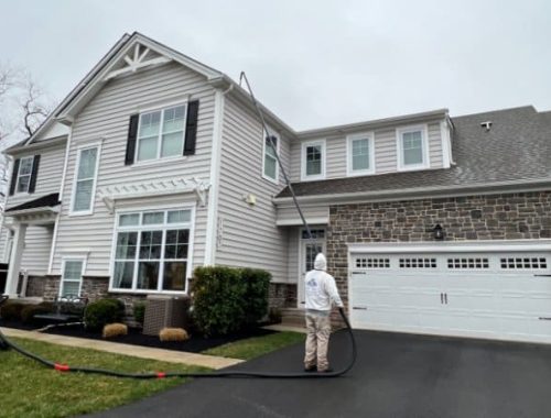 A house soft washing service cleans a two-story home with a gray exterior and stone accents. A technician in protective gear uses a long wand to apply cleaning solution to the siding.